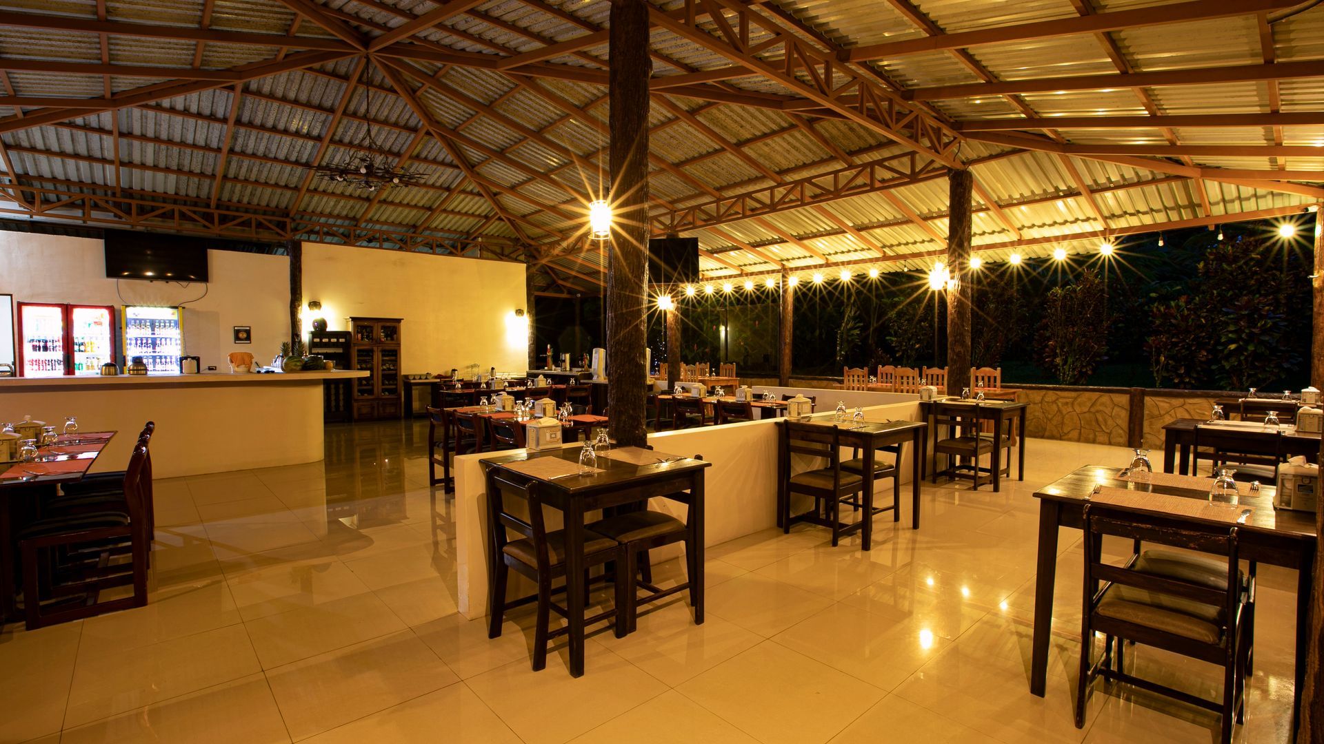 Comedor al aire libre con mesas de madera, luces cálidas y una barra a la izquierda.