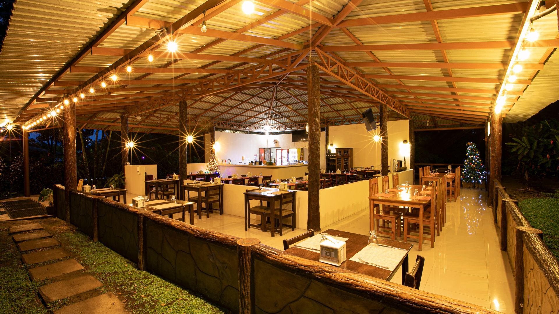 Restaurante al aire libre bajo un techo de madera con mesas, sillas e iluminación cálida por la noche.