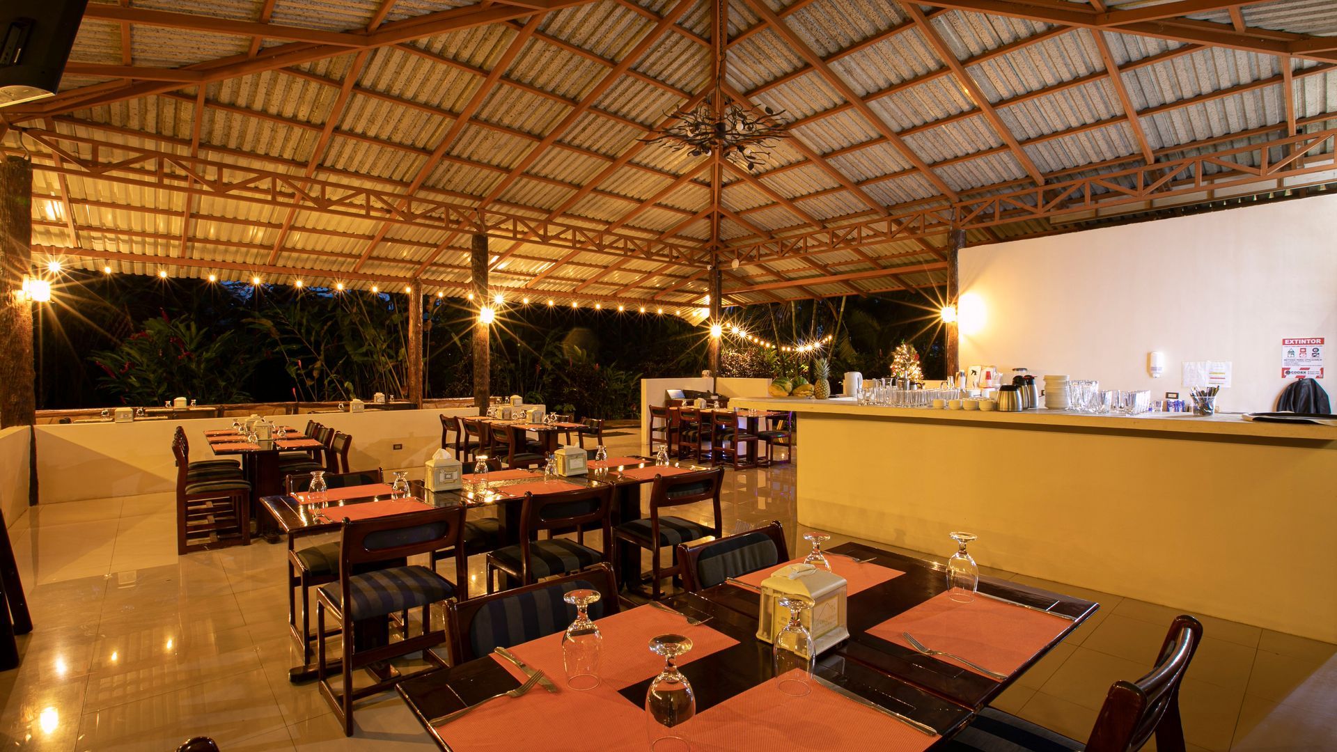 Restaurante al aire libre por la noche con mesas y sillas vacías y una barra iluminada bajo un techo de paja.