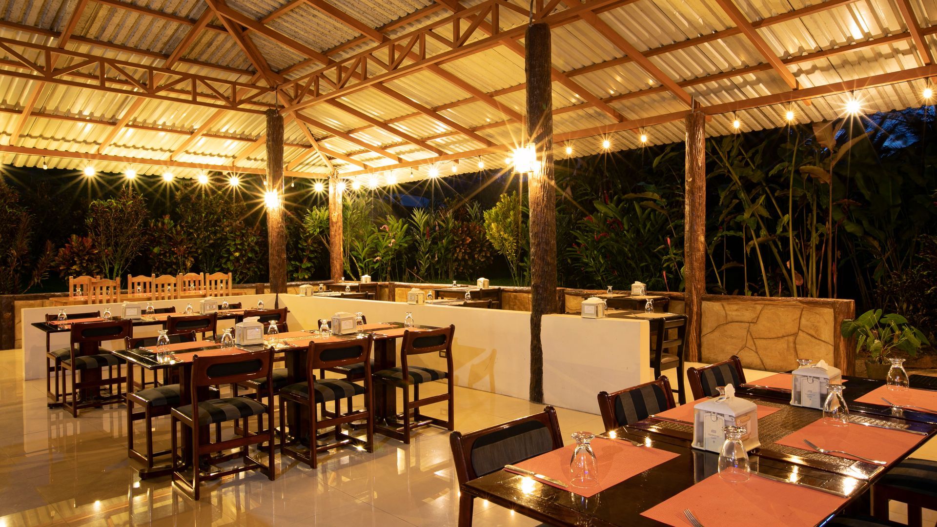 Restaurante al aire libre con mesas y sillas de madera bajo un techo de paja al atardecer.