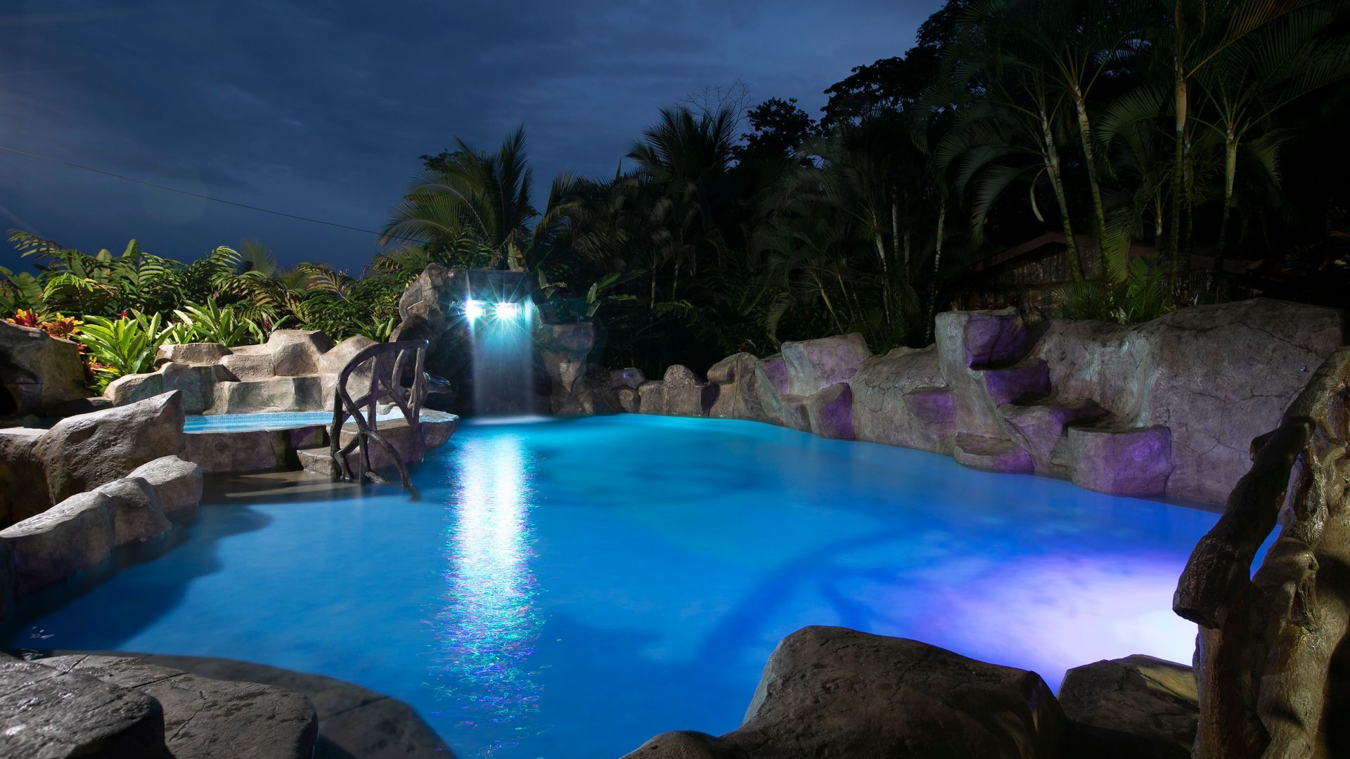 Piscina exterior iluminada con luz azul por la noche, rodeada de rocas tropicales y palmeras bajo un cielo oscuro.