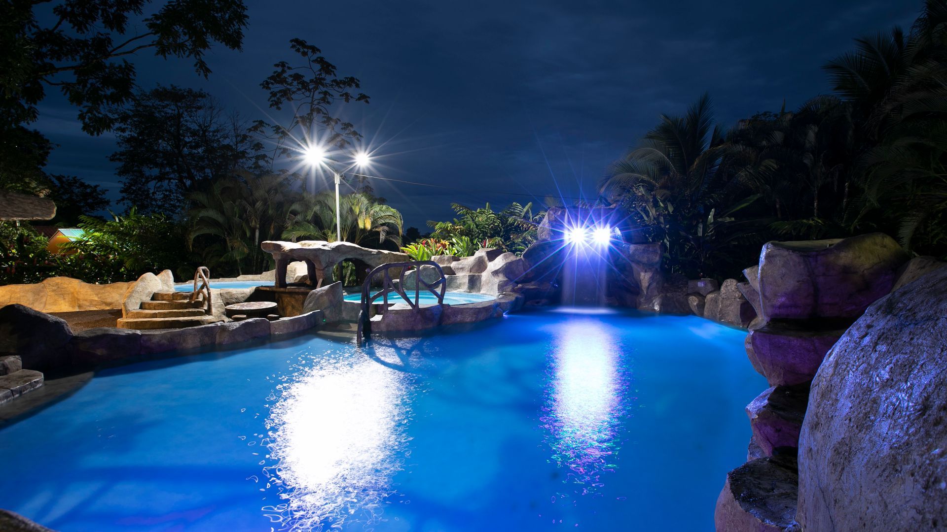 Piscina iluminada estilo resort por la noche con agua azul, palmeras y luces brillantes que se reflejan en la superficie.