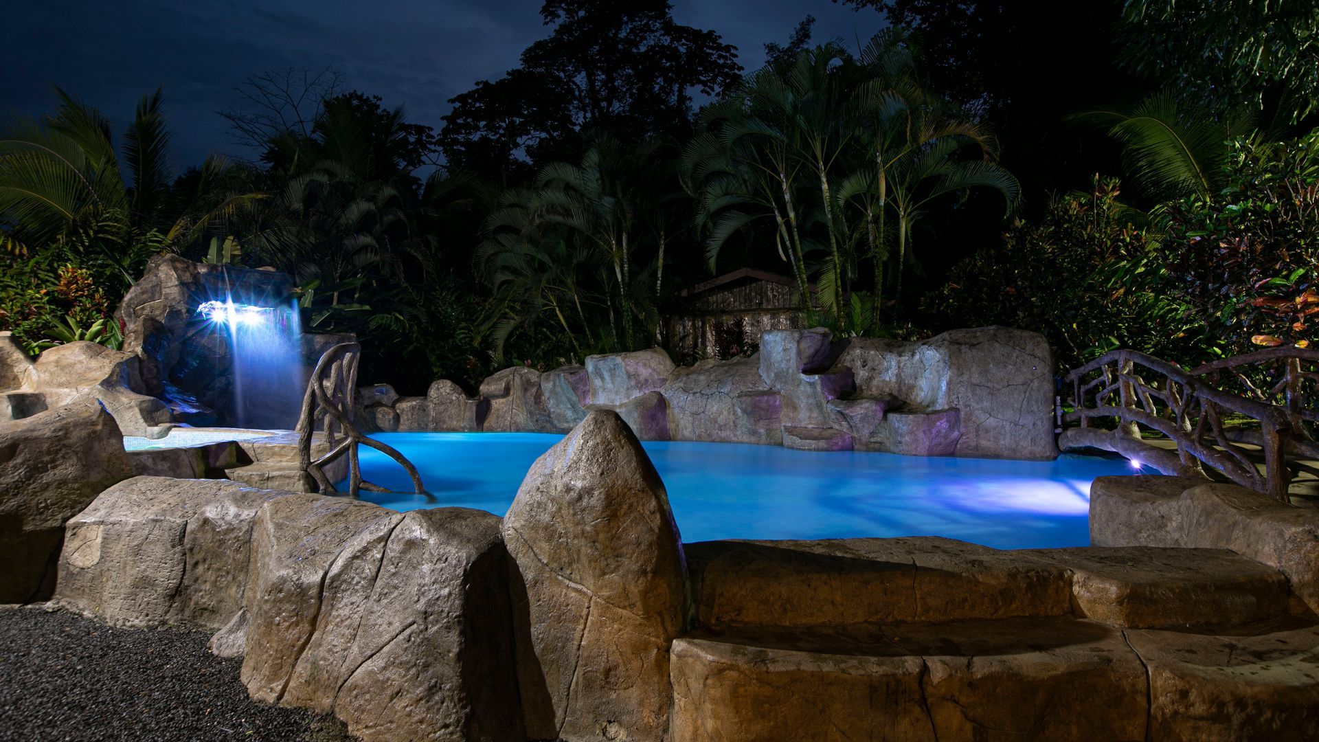 Piscina iluminada en el patio trasero con cascada de roca y paisajismo tropical por la noche.