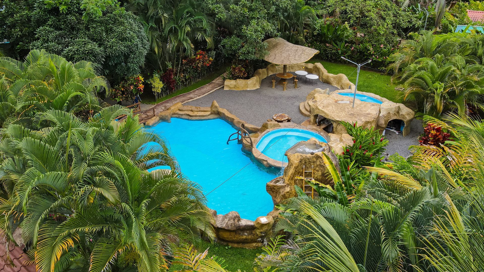 Una piscina de estilo tropical con agua azul brillante, bordes rocosos, palmeras y exuberante vegetación.