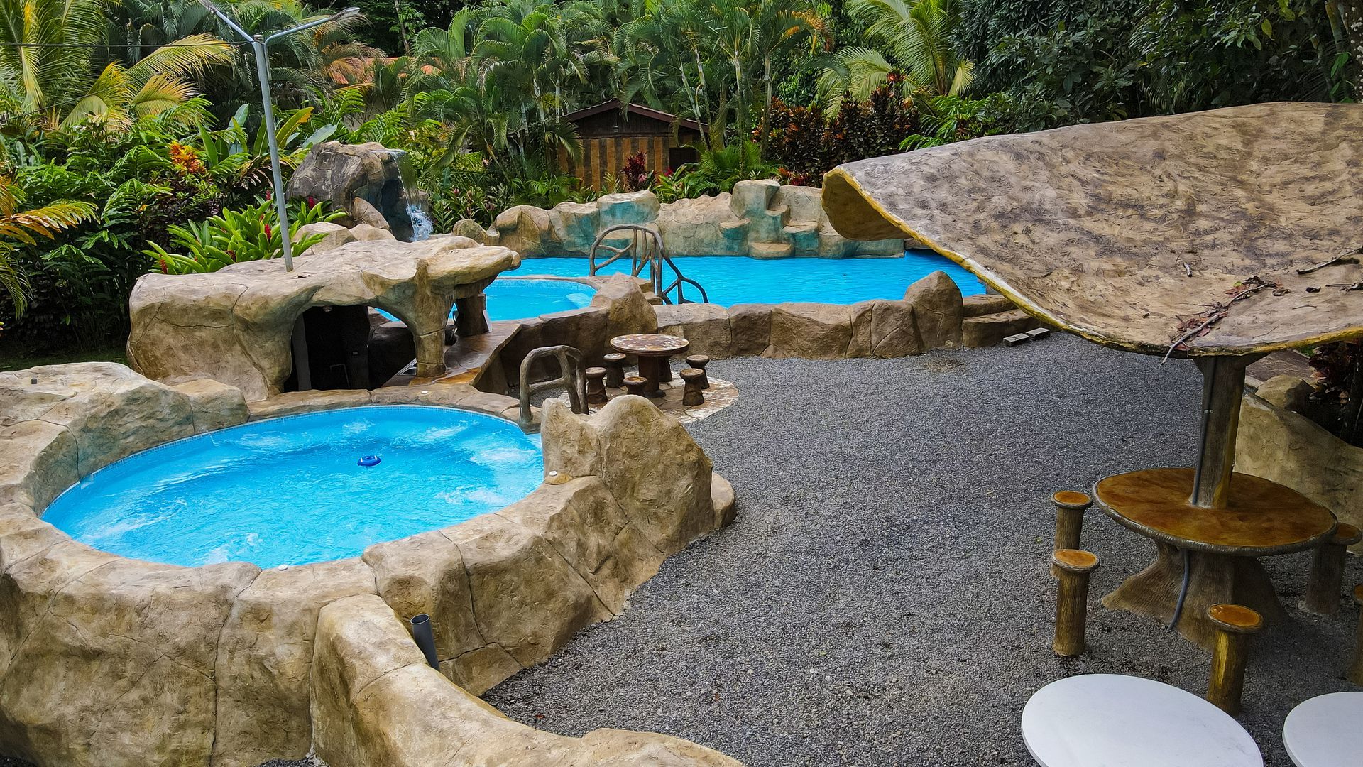 Piscina rocosa de un complejo turístico con agua azul brillante, sombrillas de paja y exuberante vegetación tropical.