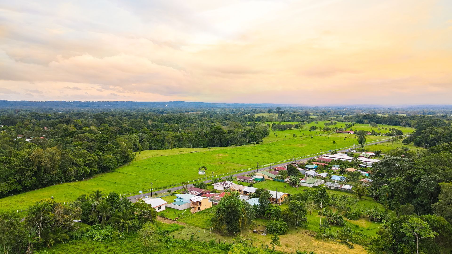 Vista aérea de un pueblo rural rodeado de exuberantes campos verdes y bosques bajo un cielo de atardecer en tonos pastel.