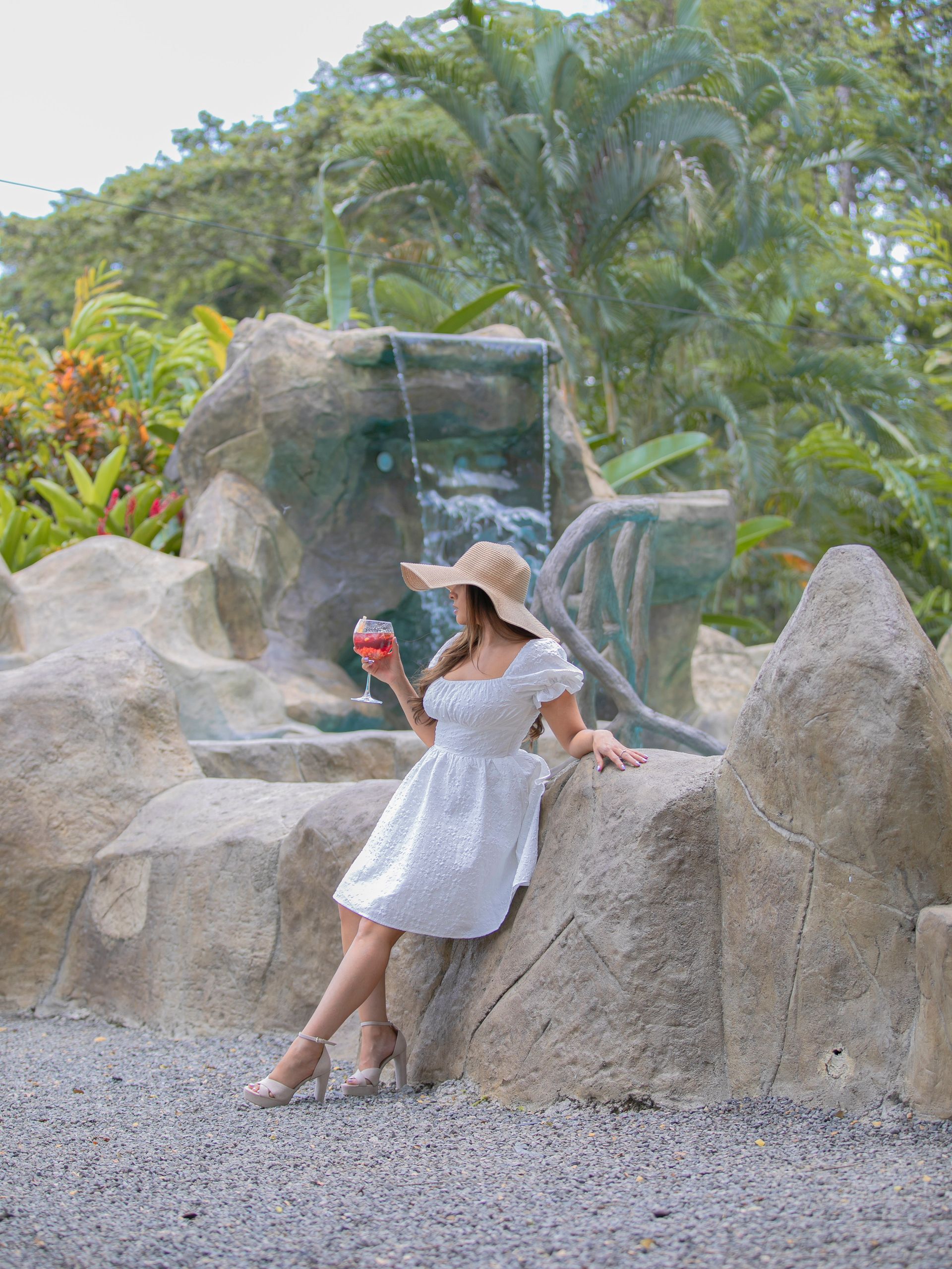 Mujer con vestido y sombrero blancos posando sobre una roca junto a una cascada en un jardín tropical.