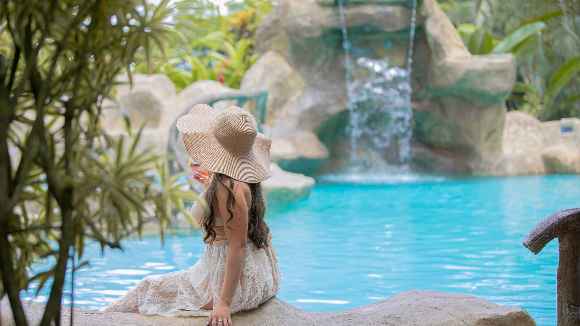Una mujer con sombrero de paja está sentada junto a una piscina de color turquesa con cascada y vegetación tropical.
