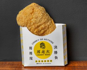 Large, breaded fried chicken cutlet in a branded paper container, on a wooden surface against a dark gray background.
