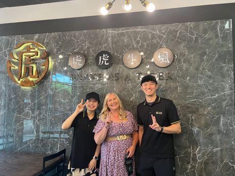 Three people pose in front of a wall with Chinese characters. A woman in a floral dress stands between two men.