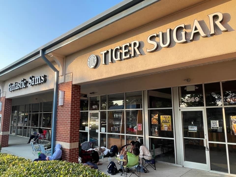 Exterior of Tiger Sugar boba shop with people waiting outside; tan building, large windows.
