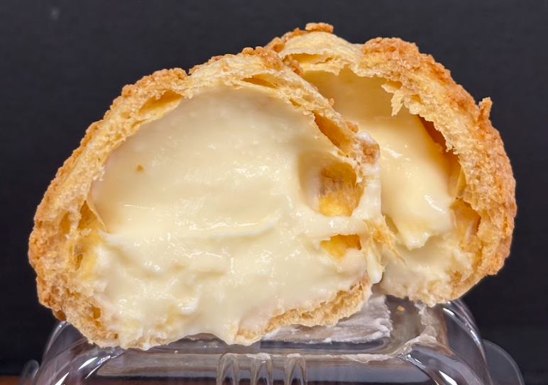 Cream puff cut in half, revealing creamy custard filling. Golden brown pastry, dark background.
