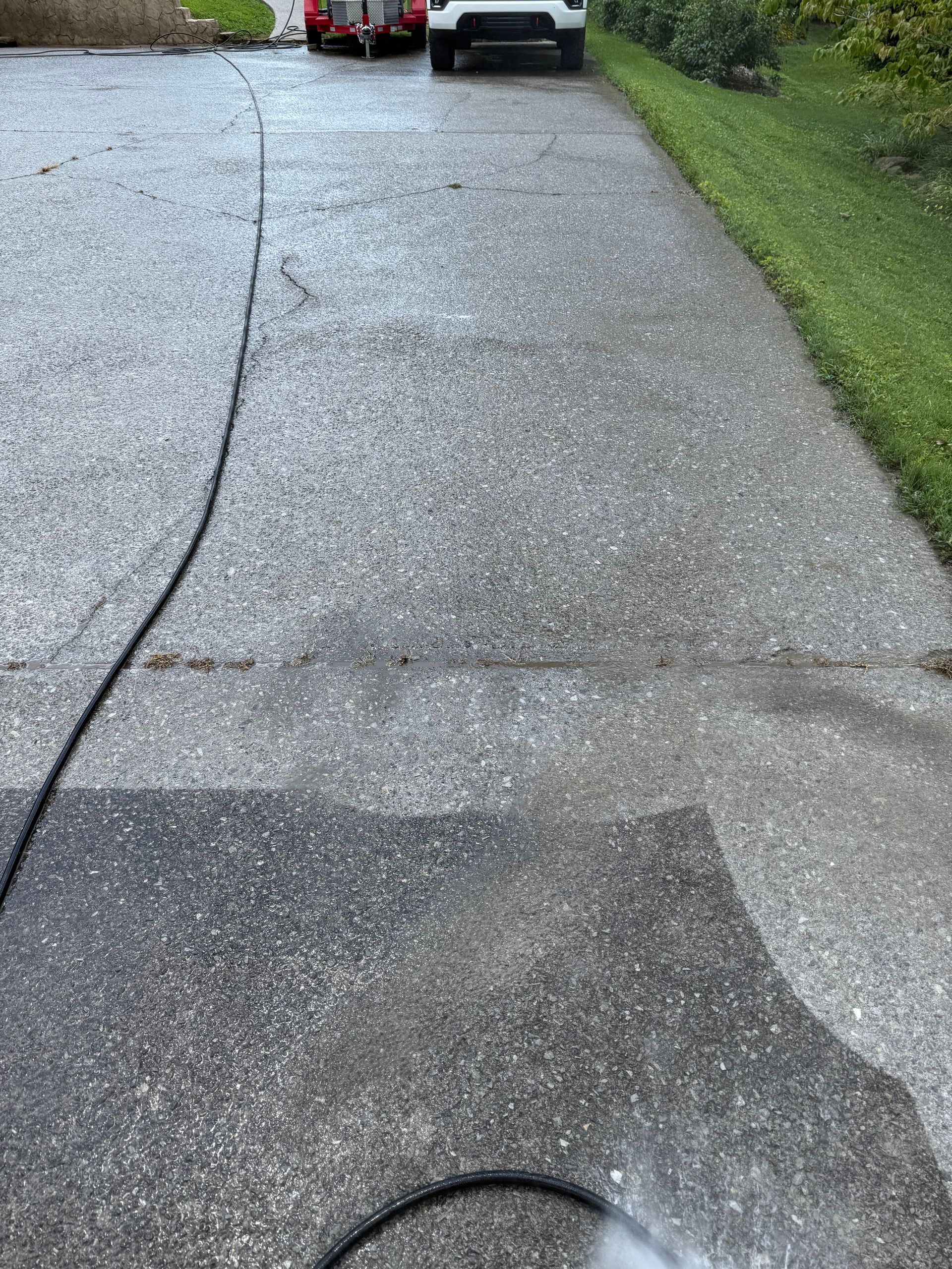 A person is power washing a wet, gray concrete driveway. Green grass borders the right side.