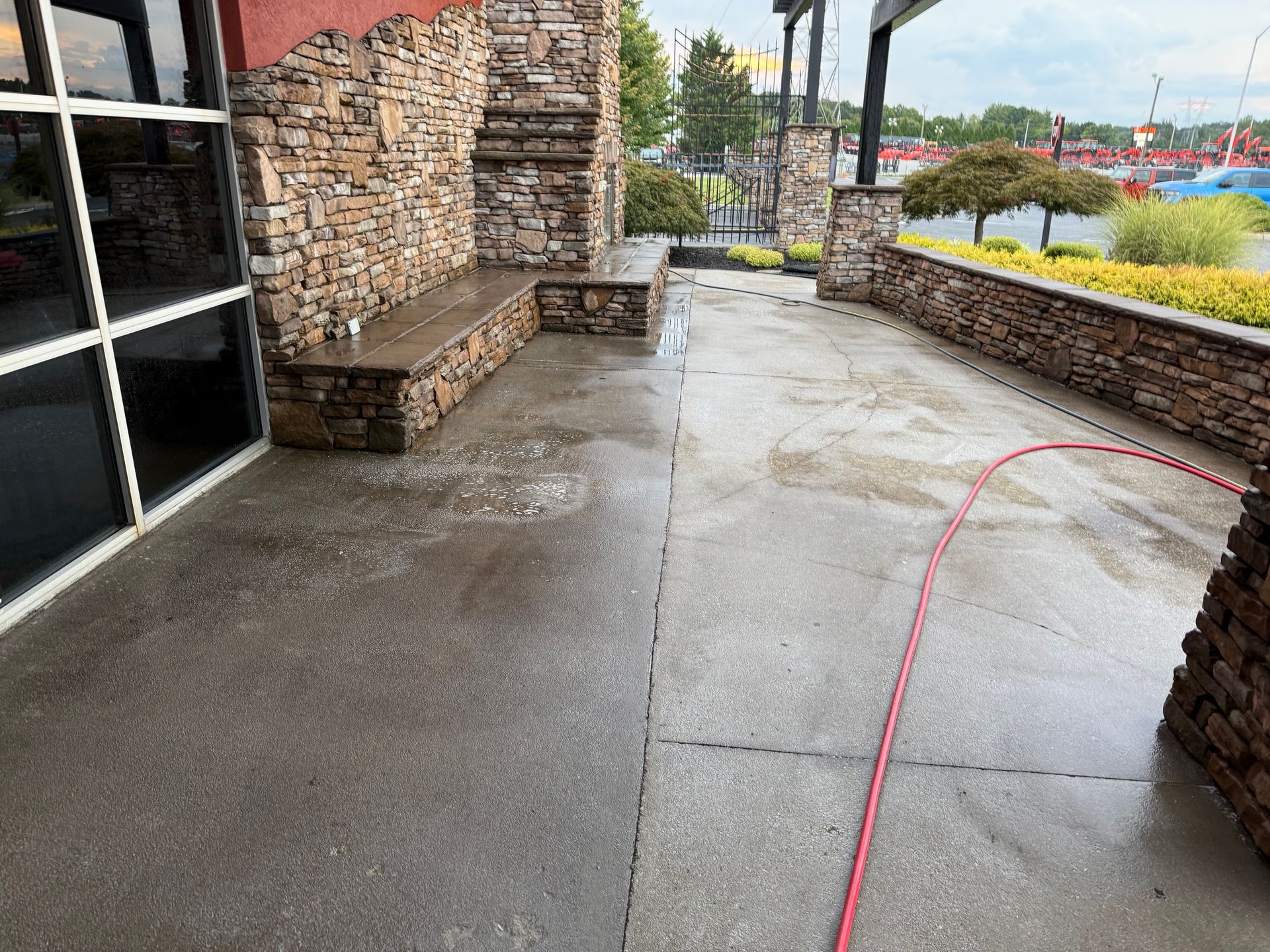A concrete patio, half cleaned with water, next to a stone wall and building with red hose.