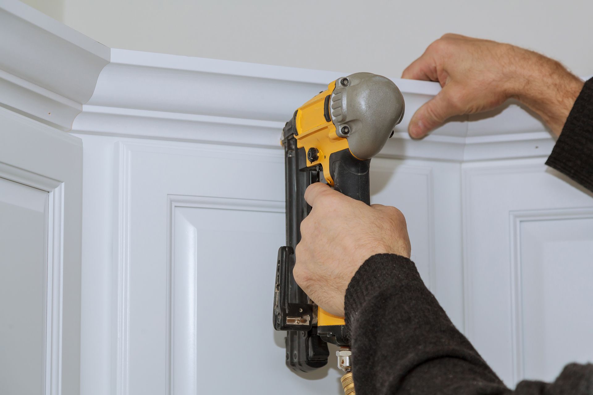 Hands using a nail gun to install white crown molding on a wall corner