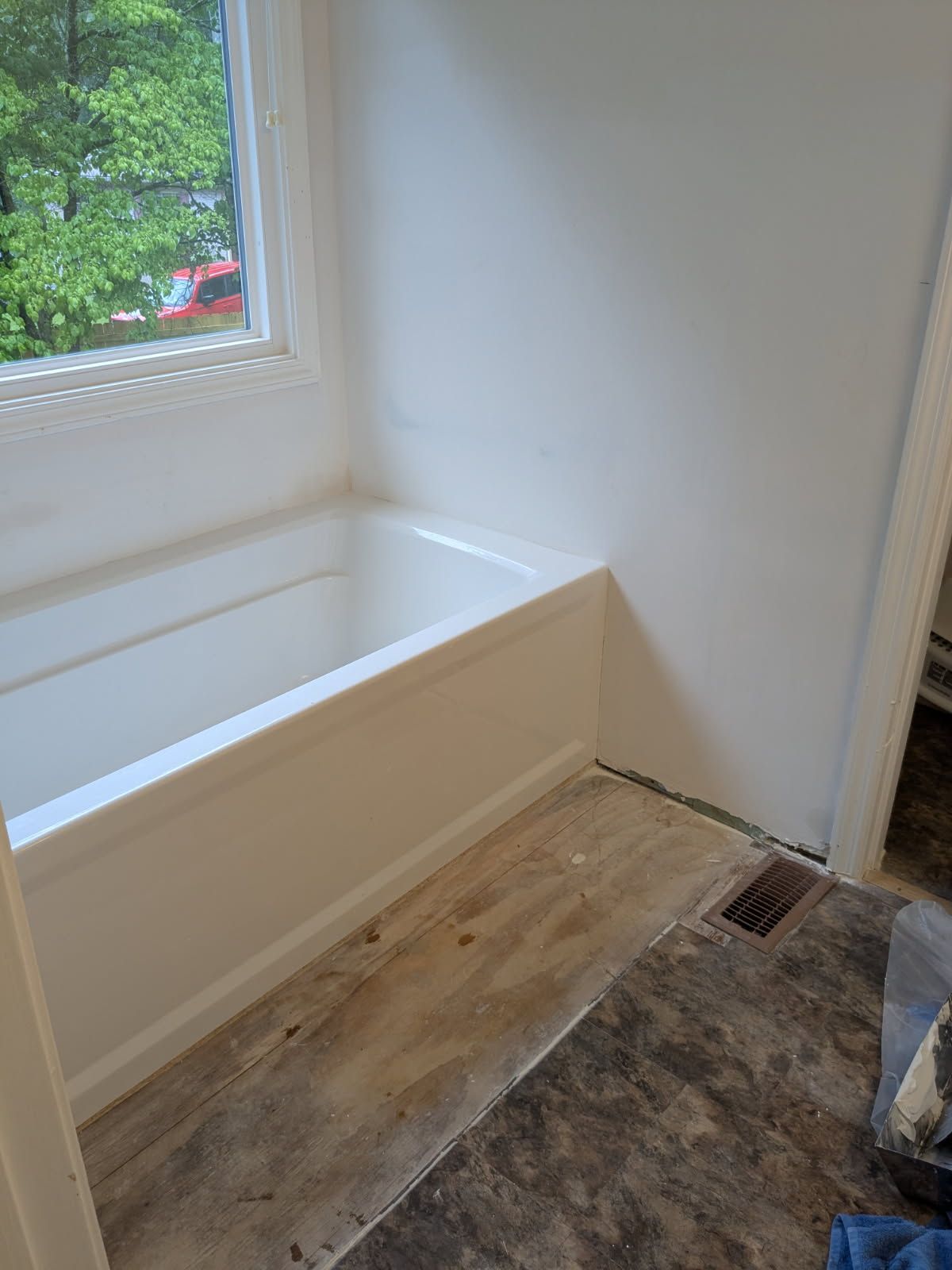 Small empty bathroom with a white tub, window, and unfinished floor near a doorway