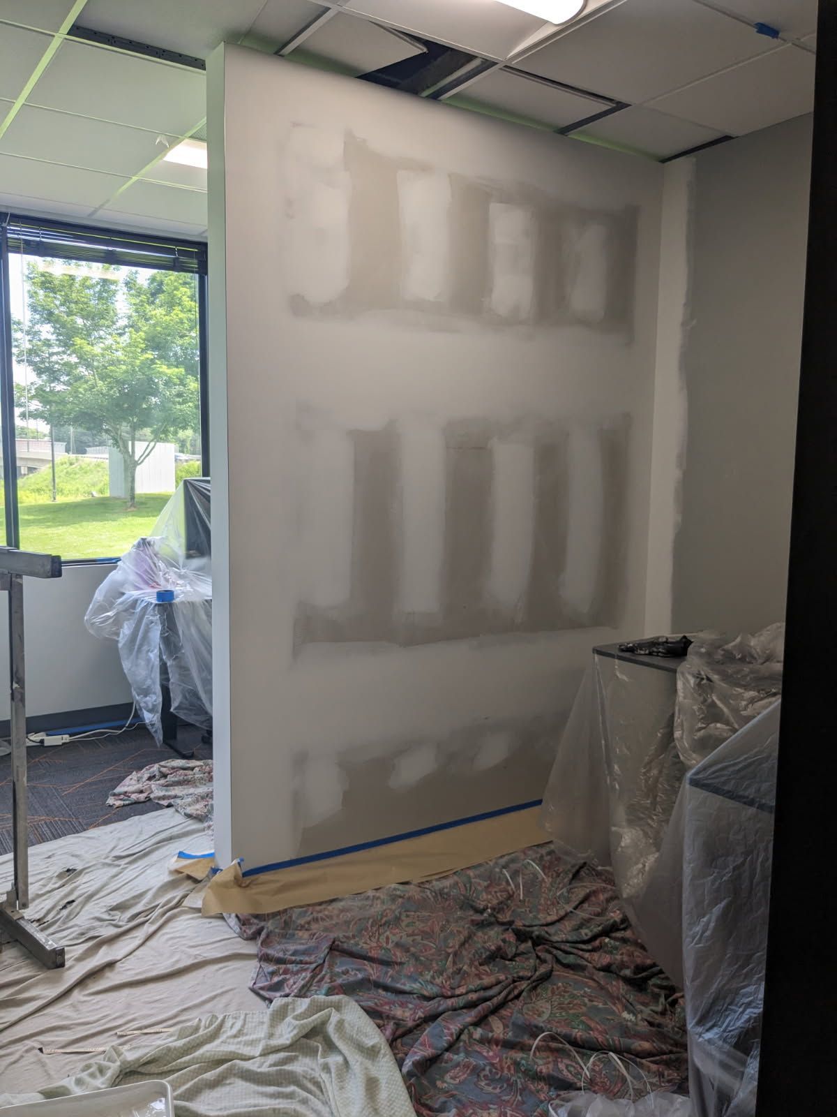 Partially drywalled office wall under renovation, with plastic-covered furniture and debris on the floor