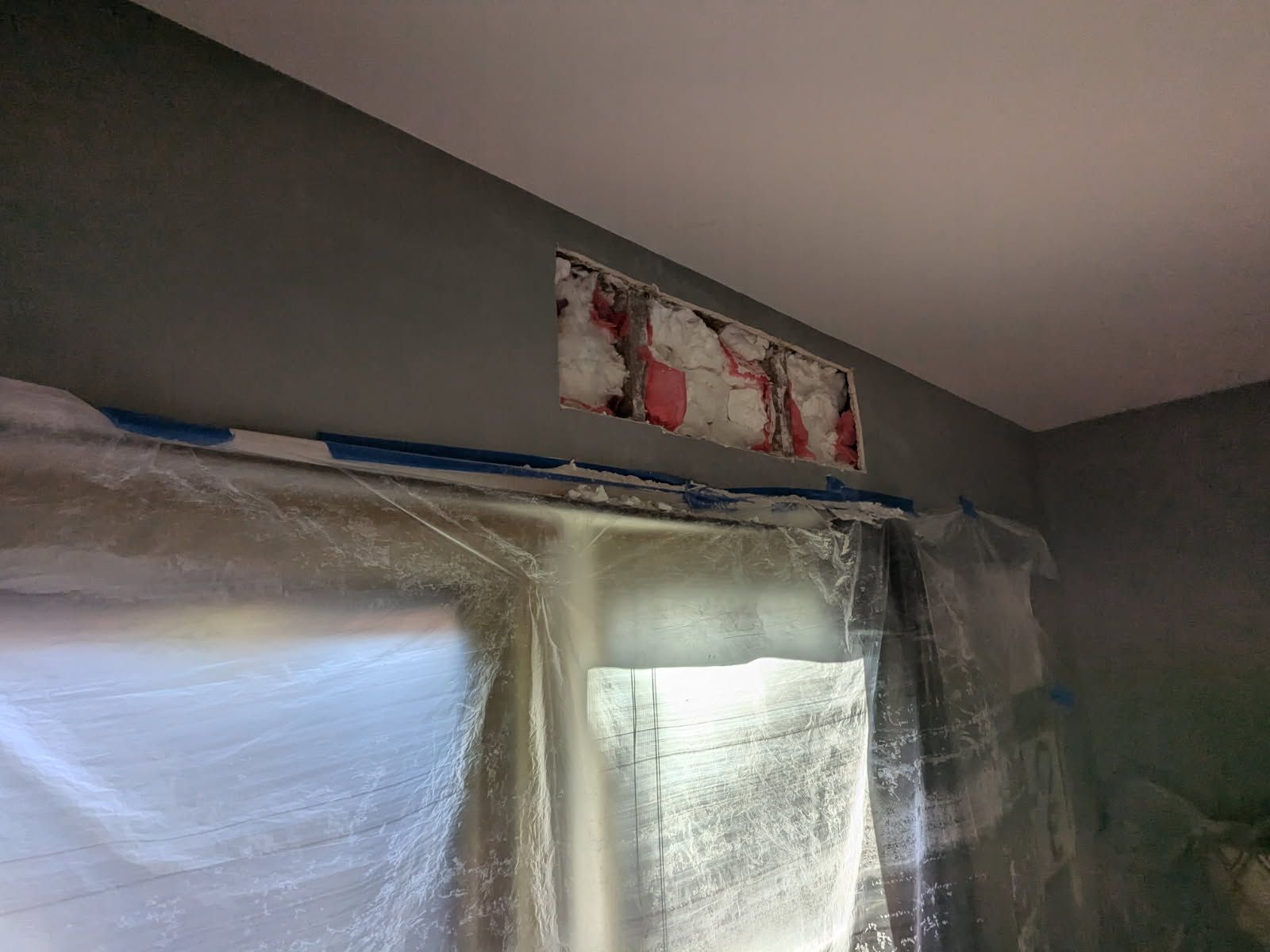Plastic-covered windows in a room with a damaged patch near the ceiling.