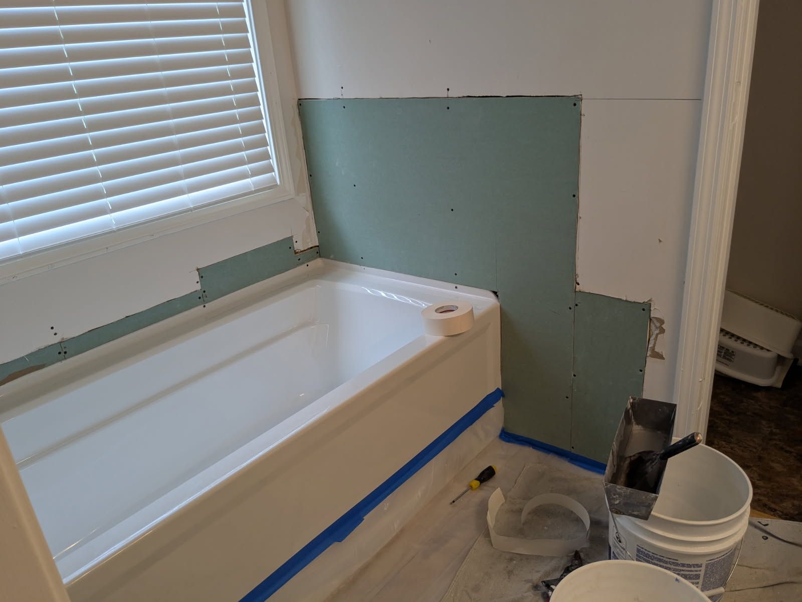 Bathroom under renovation with a white tub, green drywall patching, and paint supplies on the floor