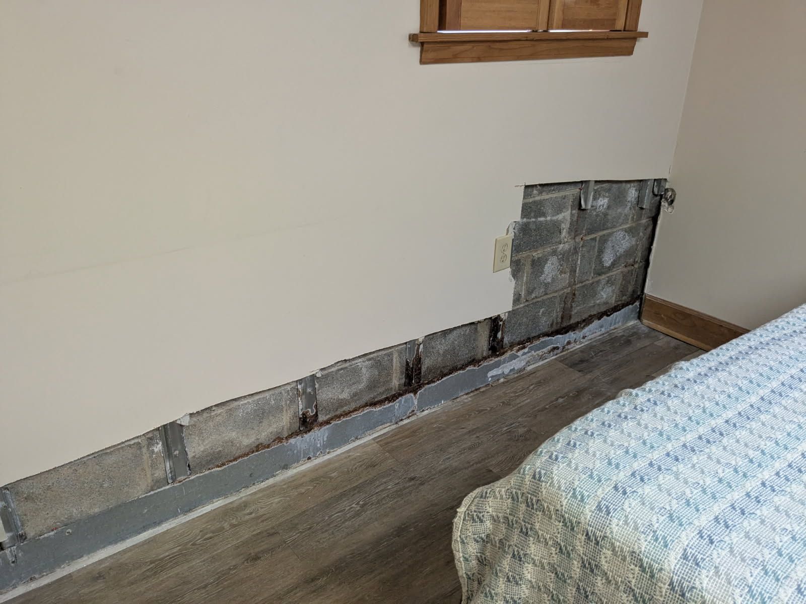 Wall with damaged drywall exposed near a bed in a bedroom.