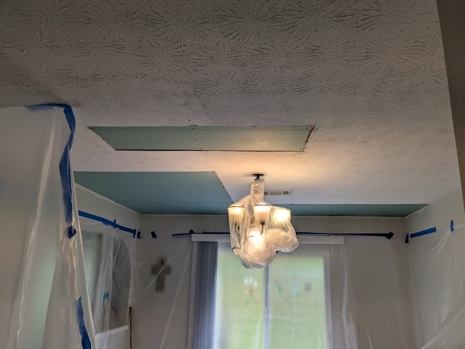 Partially covered room with plastic sheeting, blue tape, a hanging light fixture, and an open ceiling panel