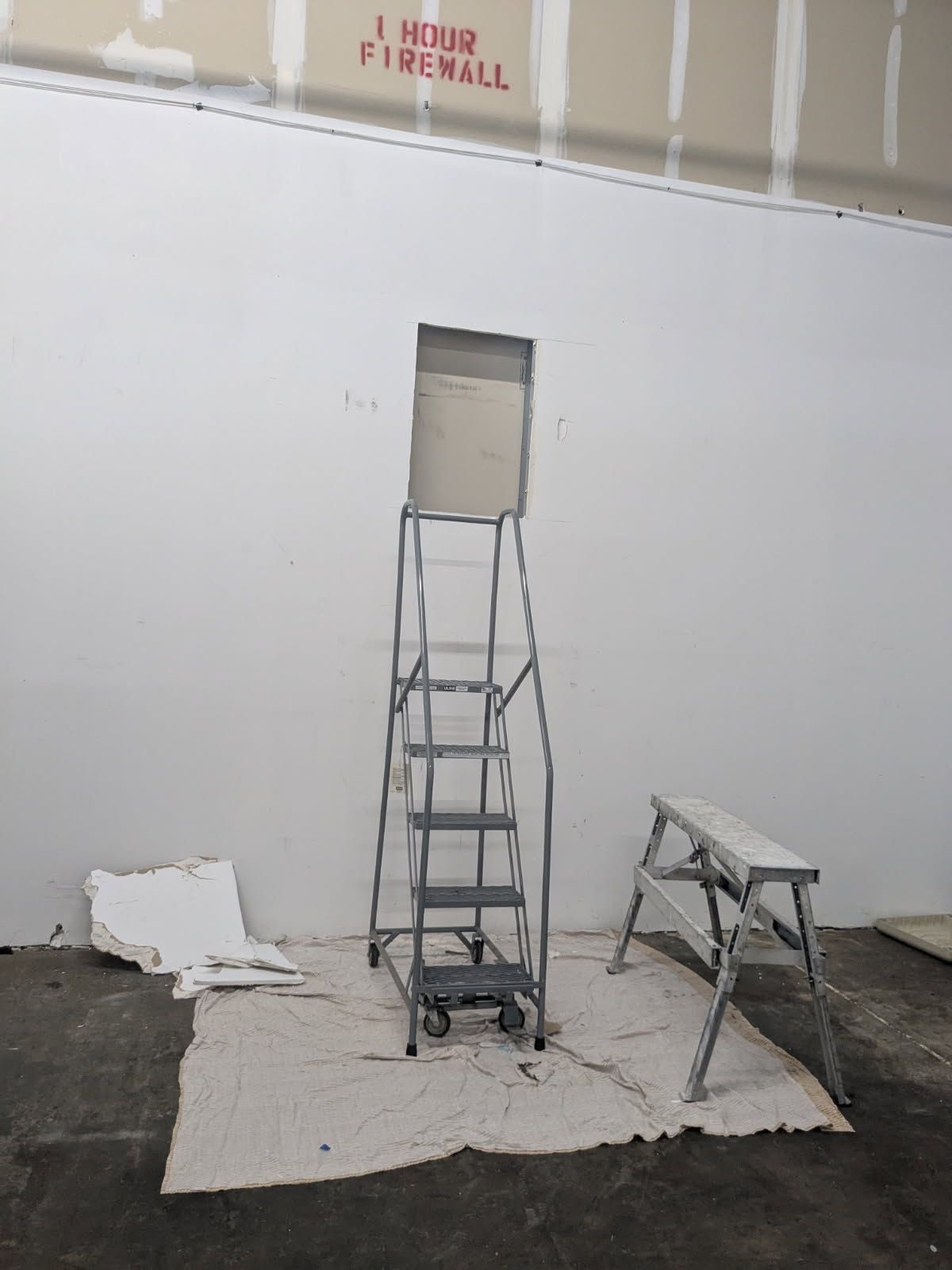 Ladder and scaffold in a room with a wall opening and protective floor covering, under fire alarm signage