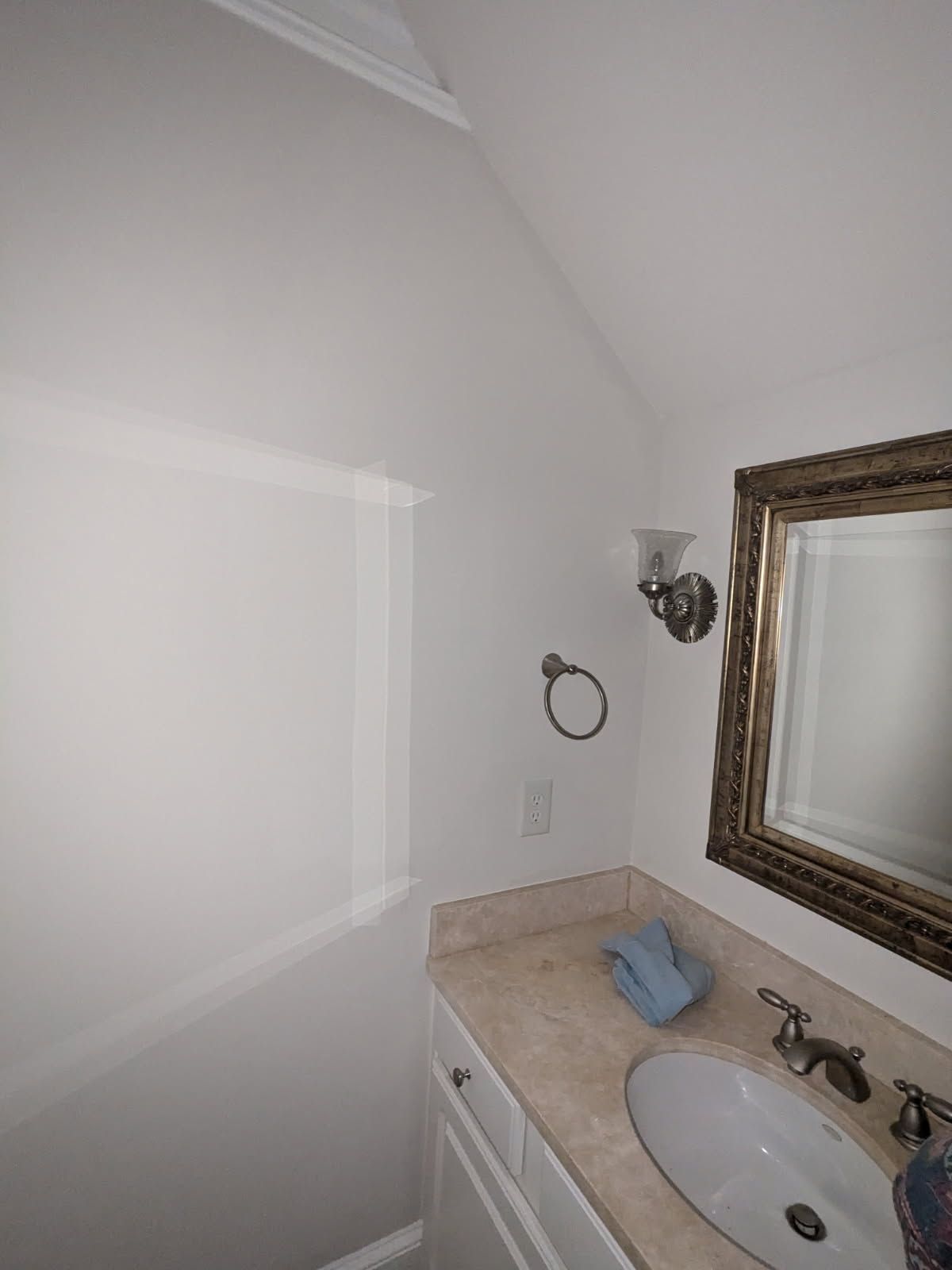 Small bathroom vanity with beige countertop, white sink, ornate mirror, wall sconce, and folded blue towel.