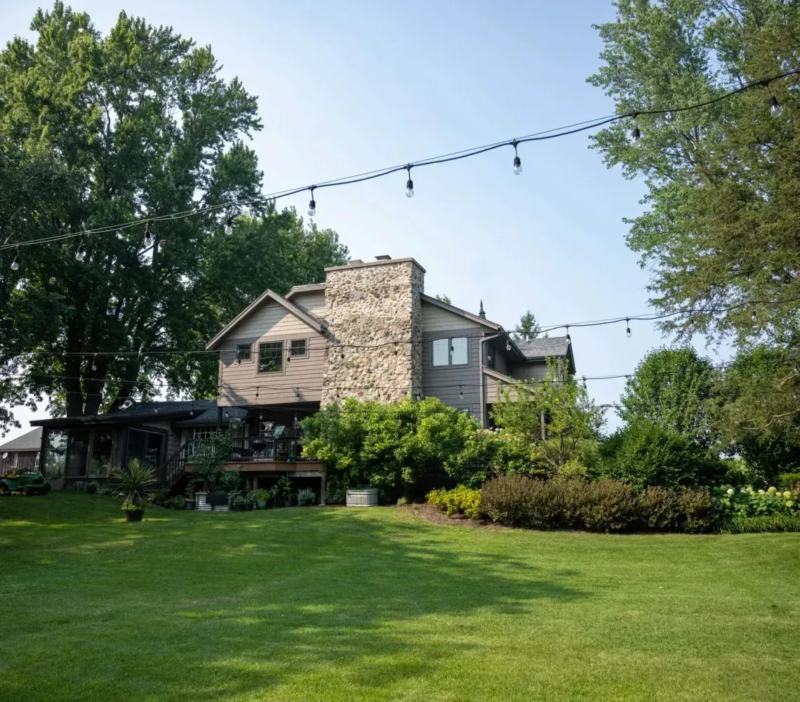 A multi-story home with a stone chimney sits behind a green lawn, framed by large trees and strings of overhead lights.