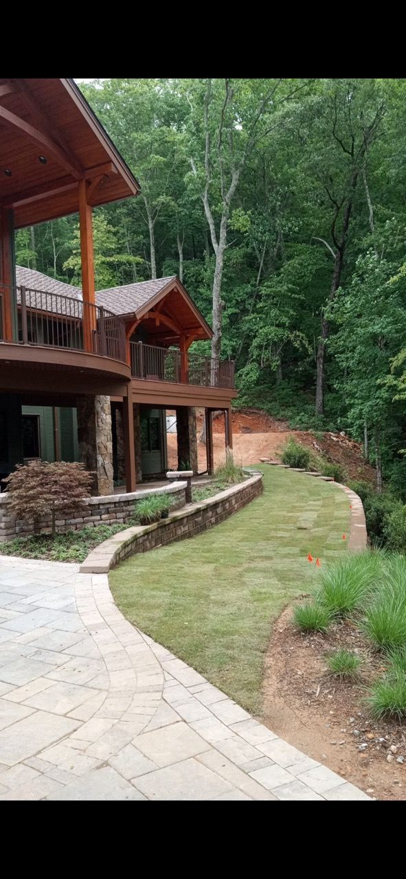 A wooden house with a stone patio and curved lawn sits beside a forest and a sloped, red-dirt hillside.