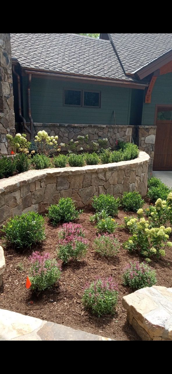 A stone retaining wall surrounds a mulched garden bed filled with small shrubs and flowering perennials near a house.