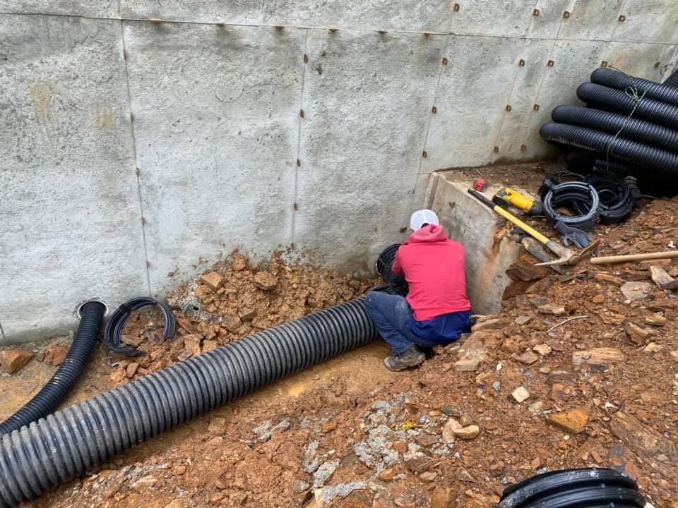 Worker in red jacket installing black drainage pipe beside a concrete wall at a muddy construction site