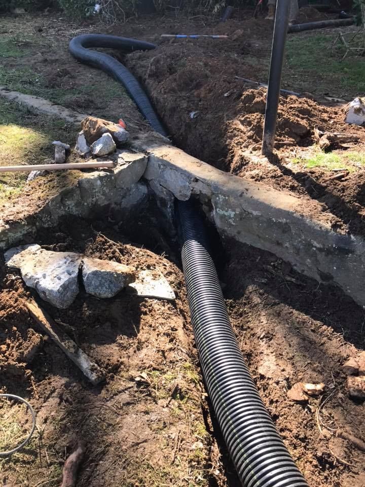 Black drainage pipe running through a dug trench under a concrete slab in a dirt yard.