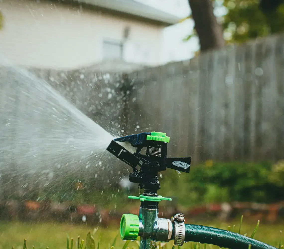 A green and black oscillating lawn sprinkler spraying water over grass in a backyard with a fence and house in the background