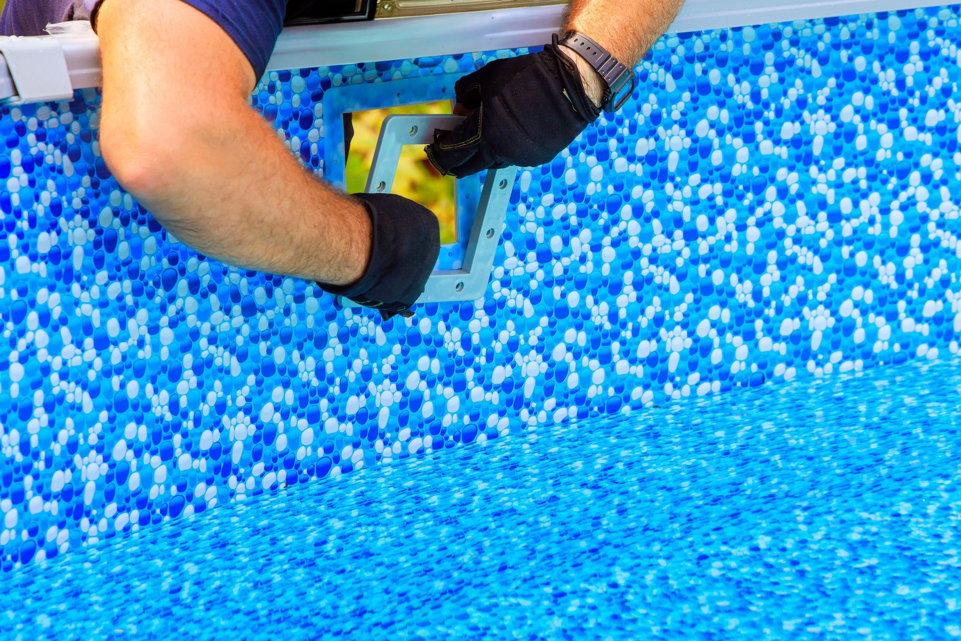 A person wearing black gloves installs a square pool skimmer faceplate onto a blue patterned pool liner.
