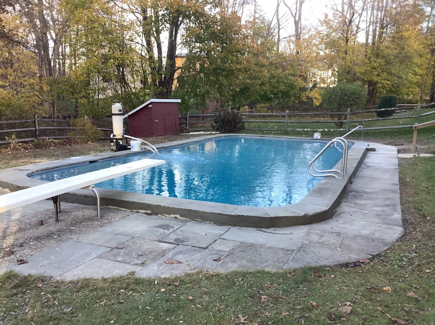 An in-ground backyard swimming pool with a diving board, surrounded by a stone patio and autumn trees in the background.