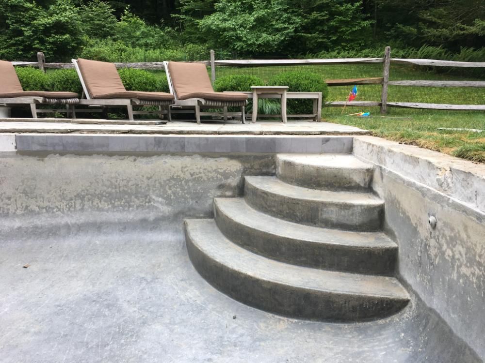 A dry, concrete swimming pool featuring a set of rounded, multi-level stairs leading to a patio with lounge chairs.