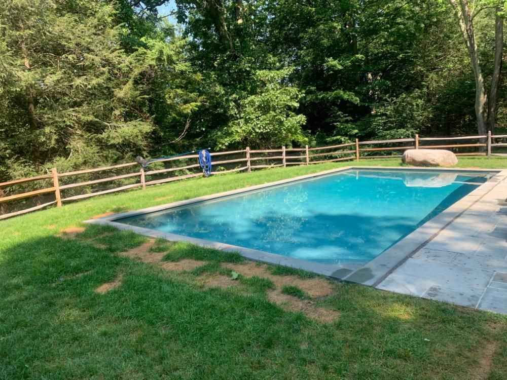 A rectangular swimming pool with blue water sits in a green lawn bordered by a wooden split-rail fence and forest.