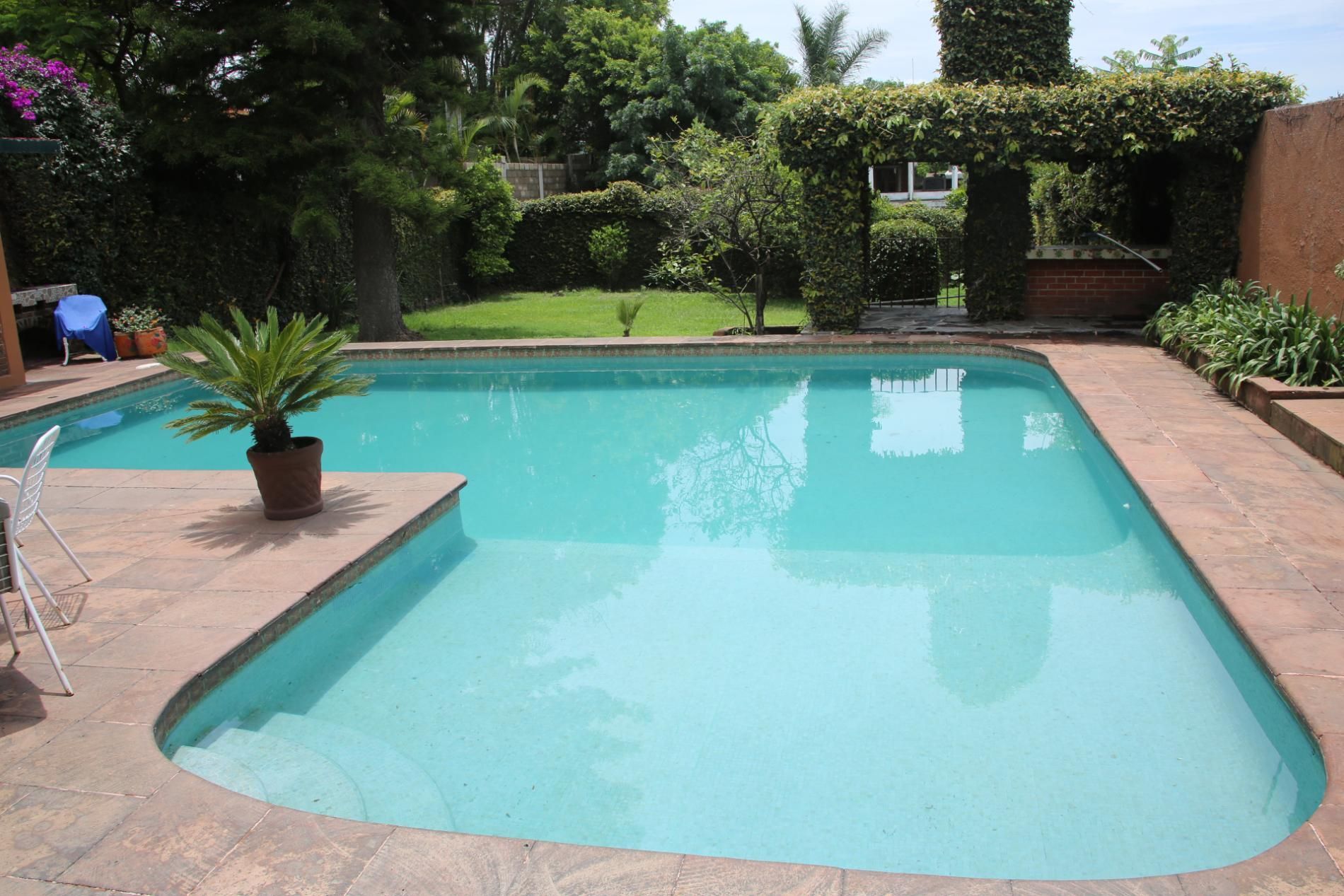 An L-shaped swimming pool in a sunny garden with stone decking, a potted plant, and a shaded seating area in the back.