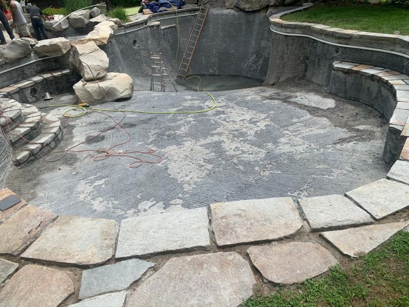 An unfinished swimming pool under construction, featuring gray stone decking, tiered stone steps, and rocky accents.