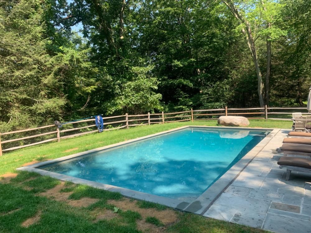 A rectangular swimming pool with blue water sits in a grassy yard beside a stone patio and a wooden split-rail fence.