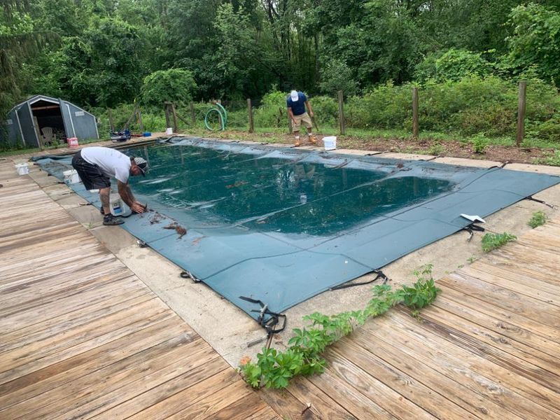 Two people work on securing a dark green swimming pool cover over an in-ground pool on a wooden deck.
