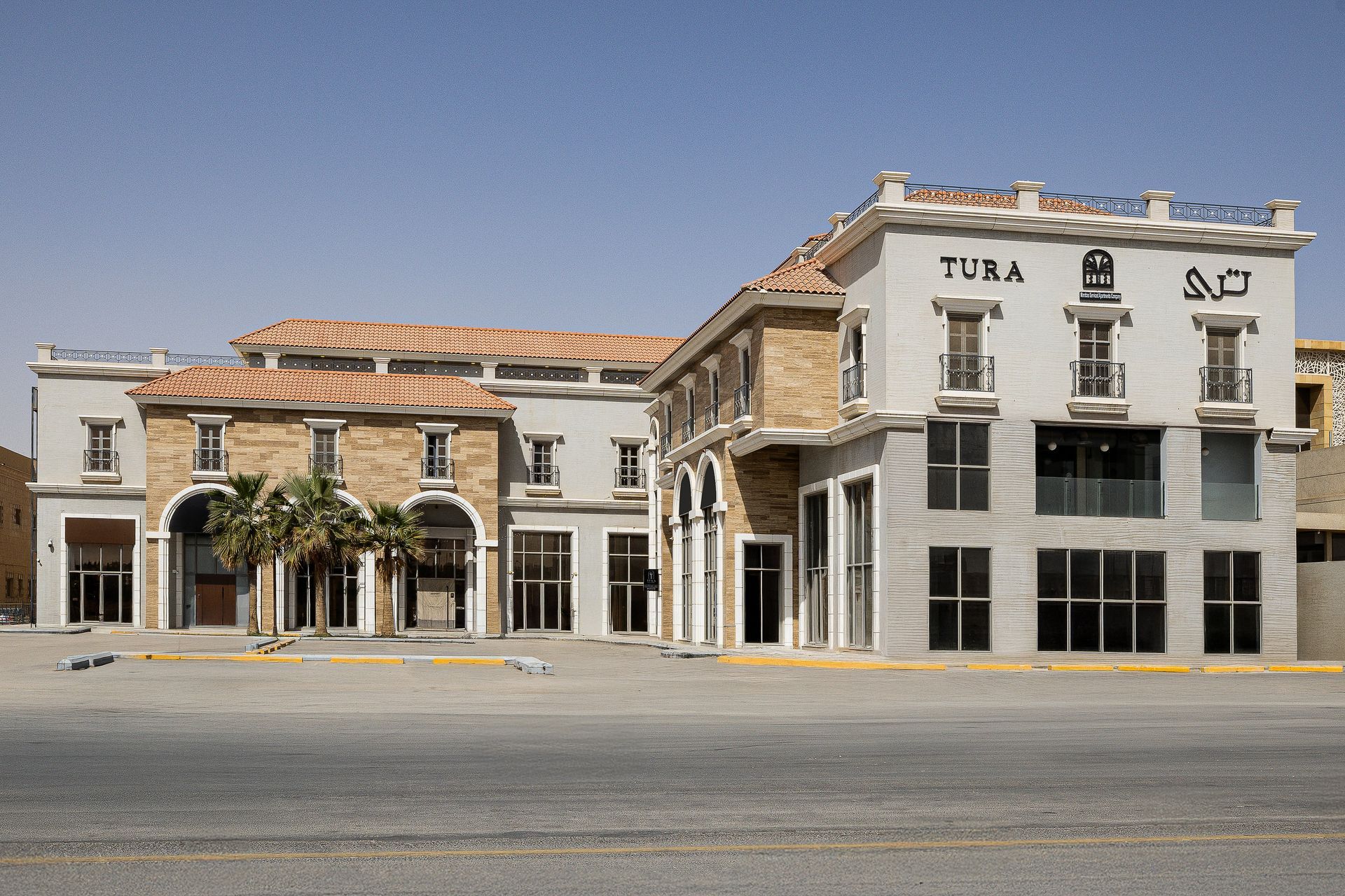 A light-colored building with Mediterranean-style architecture and a "TURA" sign sits by an empty paved parking lot.