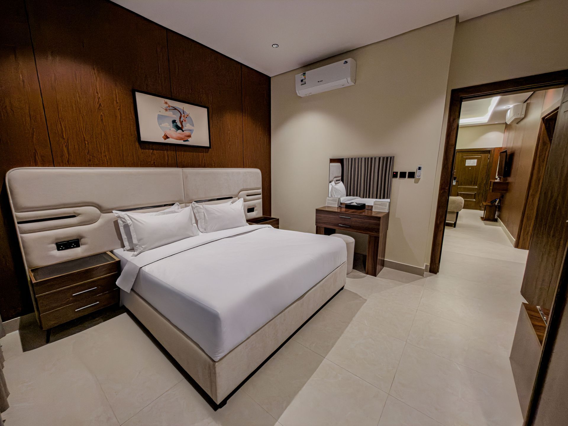 Modern hotel bedroom with a large bed, wooden feature wall, vanity, and an open doorway leading to a hallway.