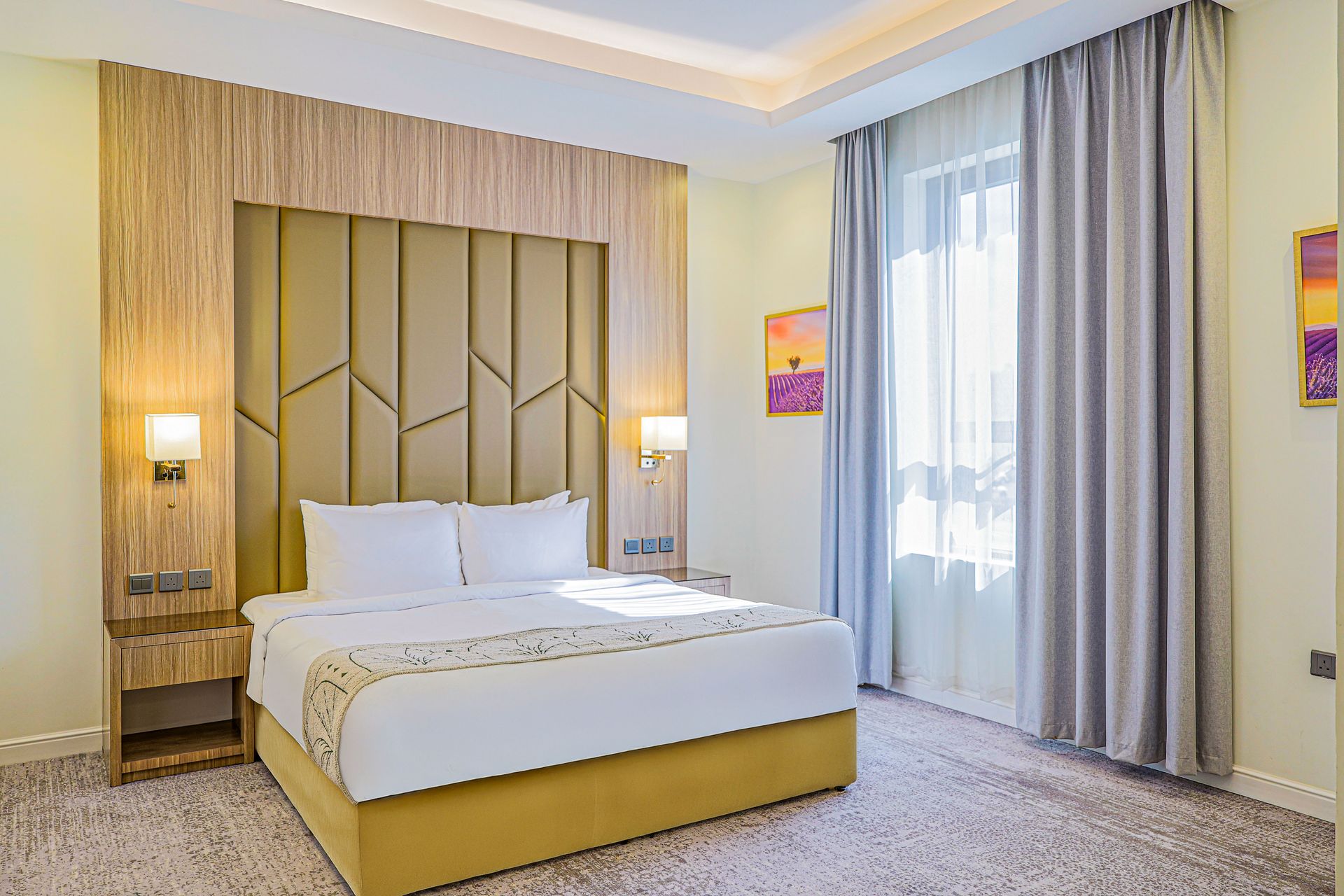 A modern hotel bedroom featuring a queen bed with white linens, a mustard yellow base, and a tan geometric headboard.