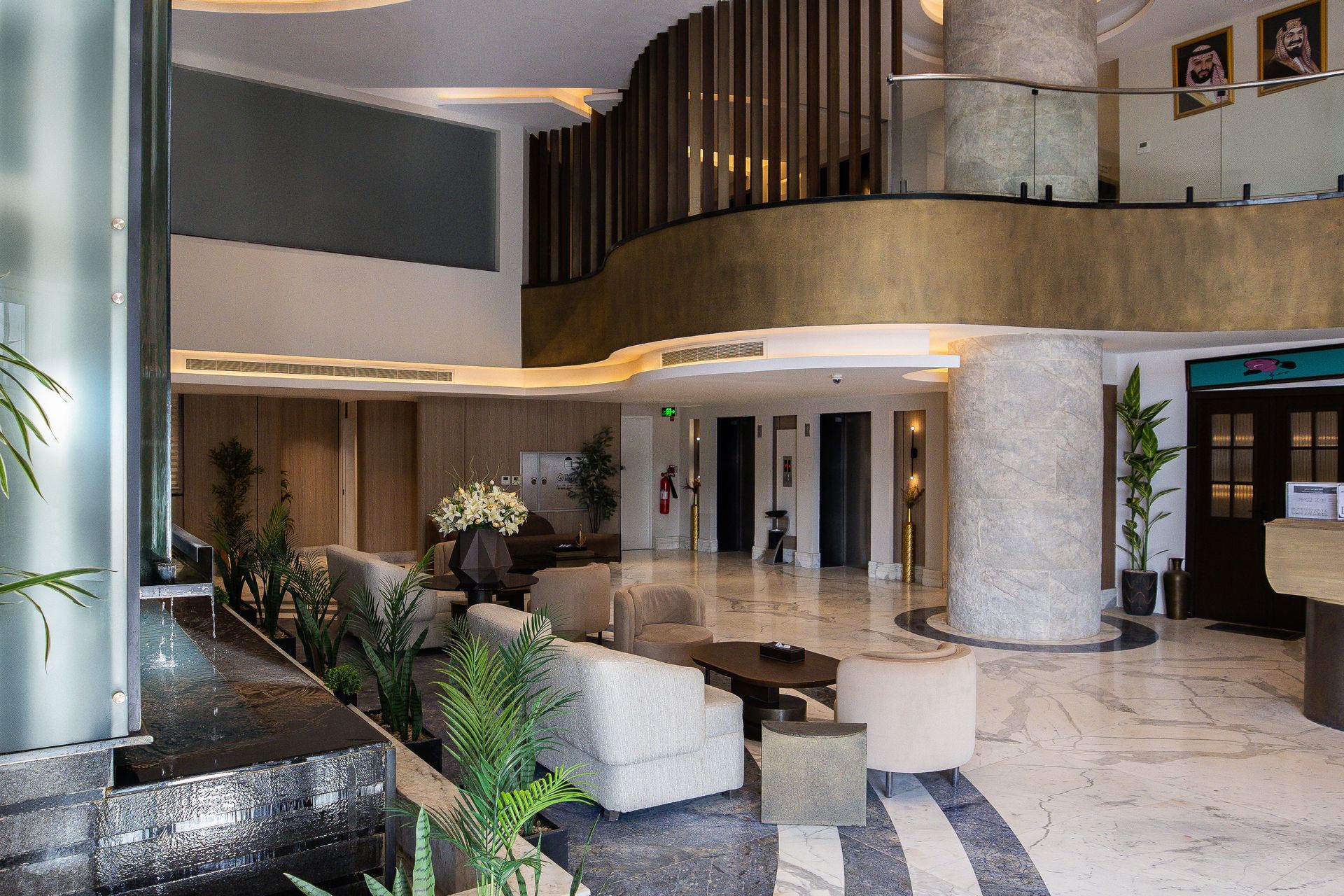 Modern hotel lobby with beige sofas, marble floors, a large central pillar, and green indoor plants under warm lighting.