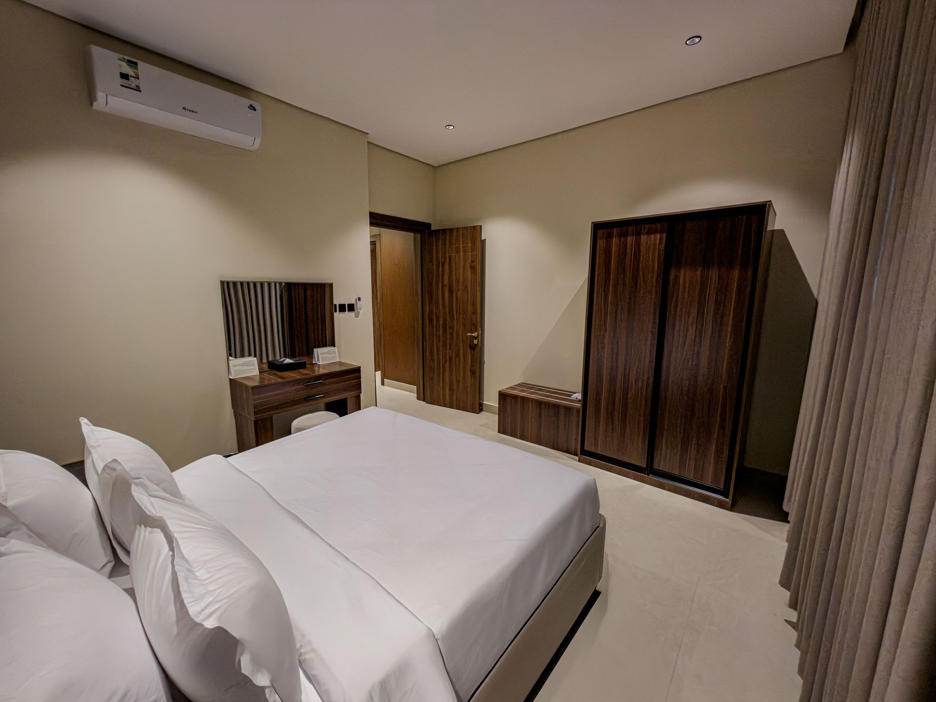 A hotel bedroom with a neatly made white bed, a wooden wardrobe, a vanity desk with a mirror, and an air conditioner.