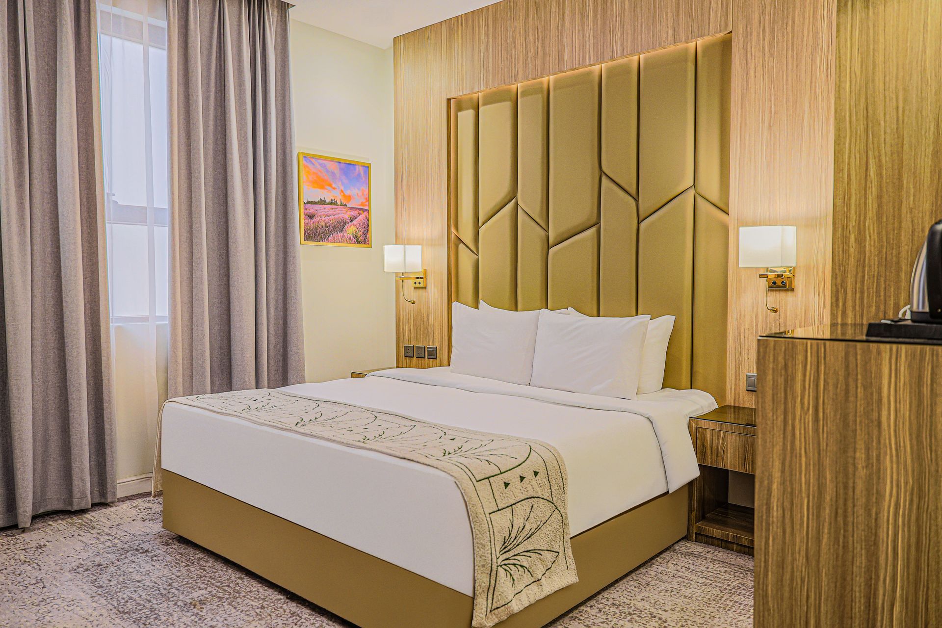 A modern hotel room with a neatly made white bed, a large gold geometric headboard, and wood-paneled walls.