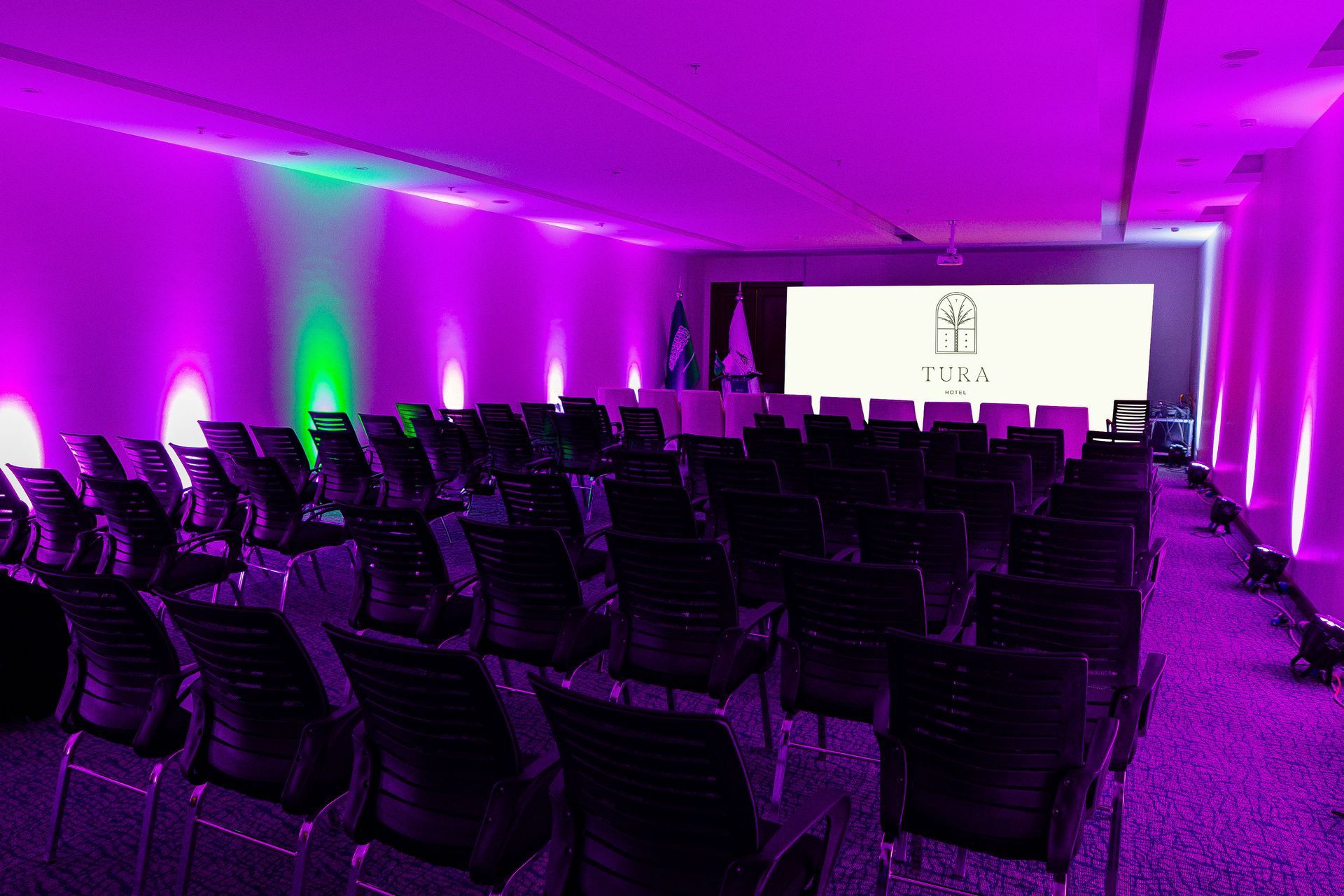 Rows of empty chairs face a large screen in a dimly lit conference room washed in vibrant purple and green lighting.