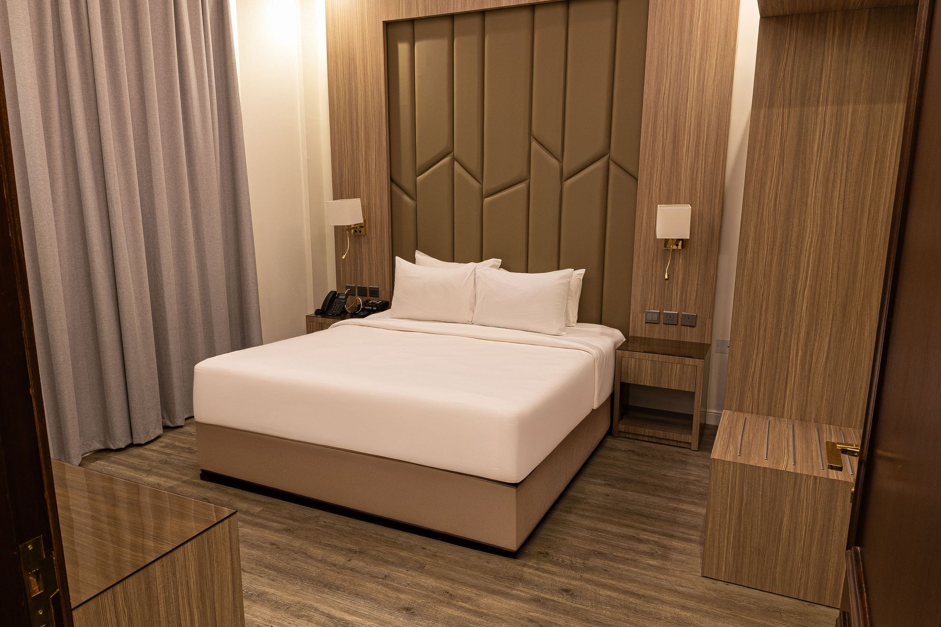Modern hotel room with a neatly made bed, wood-paneled walls, and curtains by the window.