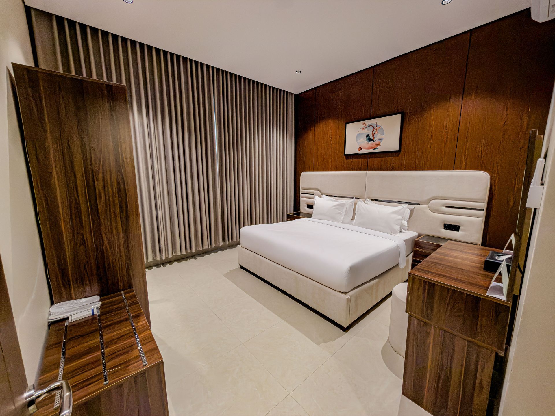 A modern hotel bedroom with a queen bed, beige headboard, dark wood accents, and floor-to-ceiling vertical striped drapes.